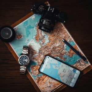 Top view of travel essentials including a map, camera, phone, watch, and lens on a wooden surface.