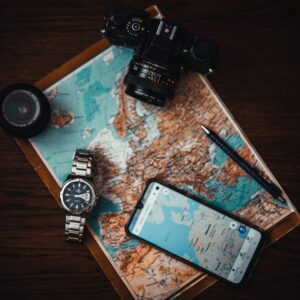 Top view of travel essentials including a map, camera, phone, watch, and lens on a wooden surface.